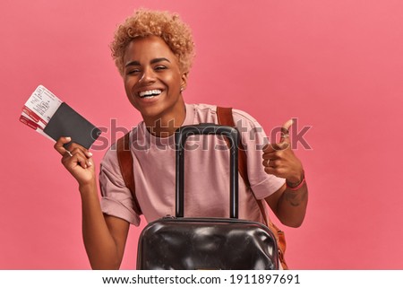 Weekends getaway. Joyful African American woman with tickets, passport and suitcase on pink background. Girl with short blond curly hair has joyful facial expression showing thumbs up recommending