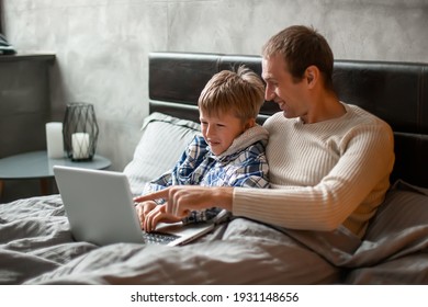 Weekends At Home. Dad And Son Lie In Bed Under A Blanket And Play On Laptop. Bachelor Party. Father And Son Are Looking For A Gift To Their Mother On March 8. Boys Shop Online.