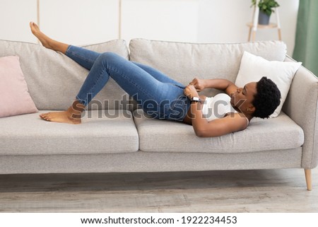 Weight Gain. Black Woman Struggling Zipping Too Small Skinny Jeans Lying On Sofa At Home. Gaining Excess Kilograms, Fat Belly And Visceral Fat Problem Concept