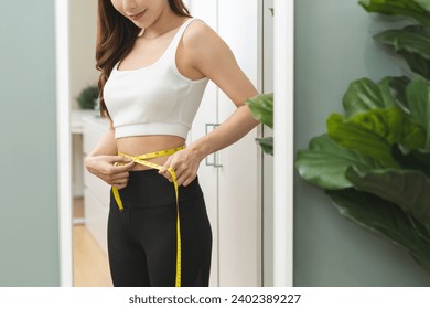 Weight lose, loss concept. Slim body asian young woman hand use tape measuring around waistline in fit sports. Girl looking reflect in mirror at home. Healthy nutrition, fitness for wellbeing beauty. - Powered by Shutterstock