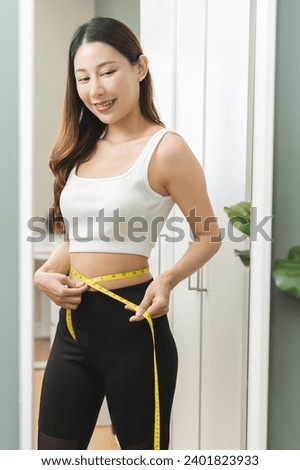 Weight lose, loss concept. Slim body asian young woman hand use tape measuring around waistline in fit sports. Girl looking reflect in mirror at home. Healthy nutrition, fitness for wellbeing beauty.