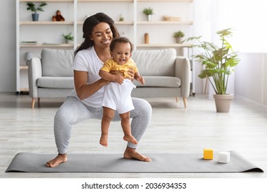 Weight Loss After Pregnancy. Happy Black Mom Training At Home With Infant Baby, Making Squats While Holding Her Cute Toddler Son In Hands, Exercising Pilates On Fitness Mat In Living Room, Copy Space