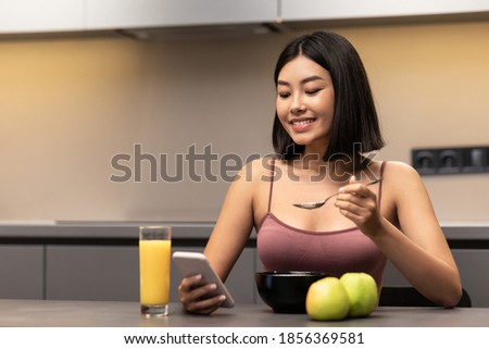 Weight Loss Application. Slim Korean Woman Using Phone Eating Oatmeal Porridge For Breakfast Sitting At Table In Modern Kitchen Indoor. Healthy Diet, Nutrition, Dieting For Slimming. Free Space
