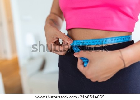 Weight Loss Concept. Portrait of millennial African American lady in top bra and leggings measuring her waist using blue tape, free copy space. Black woman checking body parameters