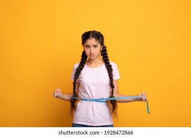 Weight Loss And Dieting. Sad And Disappointed Indian Woman Measuring Waist With Tape, Unhappy About Her Body Parameters, Standing Isolated Over Yellow Studio Background. Slimming Concept. Free Space