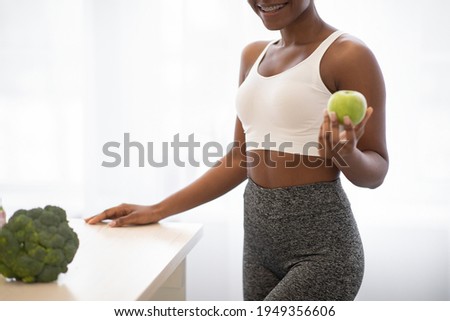 Weight Loss Nutrition. Unrecognizable African Fitness Lady Posing With Apple Standing In Kitchen Indoors, Cropped. Healthy Diet For Slimming. Cooking For Weight-Loss Concept