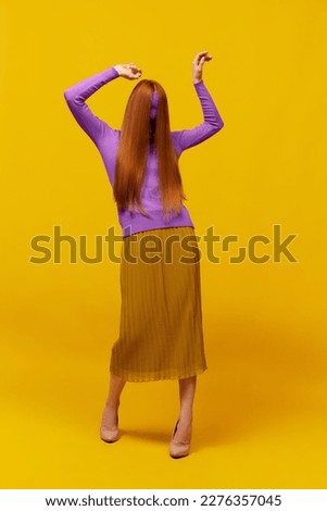 Weird dance. Faceless portrait of slim woman with long straight red hair wearing retro fashion outfit in action over yellow studio background. Impersonal emotions, body language