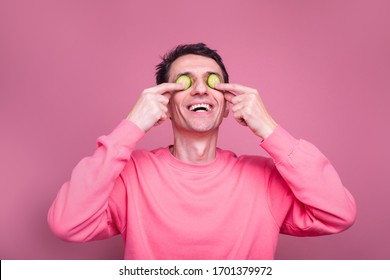 Weird Funny Young Man Hold Cucmber Slices Over Eyes And Smile Or Laugh. Stand Alone And Rtake Care Of Facial Skin. Isolated Over Pink Background