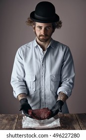 Weird Young Jewish Butcher Man With Curly Hair And Beard Wearing A Too Small Derby Hat And Faded Denim Shirt Offers A Kosher Raw Steak In His Hands Over A Wooden Table.