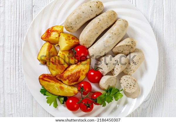 Weisswurst, bavarian white sausage of minced\
veal, pork back bacon, spices and parsley on white plate with roast\
potatoes, fresh tomatoes, horizontal view from above, flat lay,\
close-up