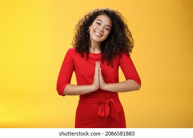 Welcome come inside. Portrait of friendly and polite good-looking female host in red dress with curly hair holding hands in namaste gesture tilting head and smiling as inviting guests in asian style