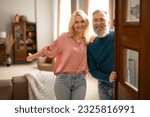 Welcome. Happy senior family couple holding opened door looking at camera, inviting guests to enter their home. Real estate owners waiting for visitor to come standing in doorway of cozy apartment