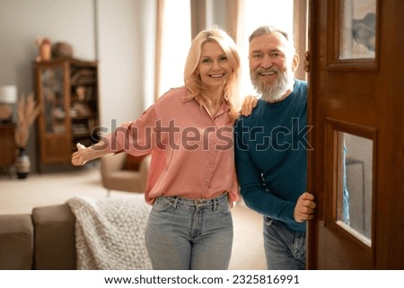 Welcome. Happy senior family couple holding opened door looking at camera, inviting guests to enter their home. Real estate owners waiting for visitor to come standing in doorway of cozy apartment