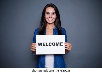 Welcome At New Home. Beautiful Modern Smiling Woman In A Jeans Shirt Is Holding A Sign With The Inscription 