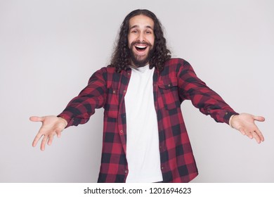 Welcome, Nice To See You. Surprised Man With Beard And Black Long Curly Hair In Casual Checkered Shirt Looking At Camera With Raised Arms And Happiness. Indoor Studio Shot, Isolated On Grey Background