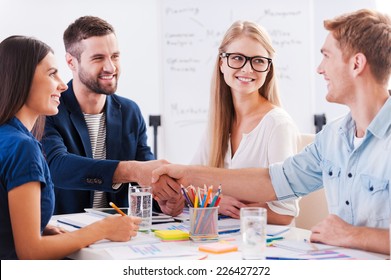 Welcome On Board! Group Of Cheerful Business People Sitting At The Table Together While Two Men Shaking Hands And Smiling 