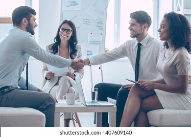 Welcome On Board! Two Handsome Men Shaking Hands With Smile While Sitting On The Couch At Office With Their Coworkers