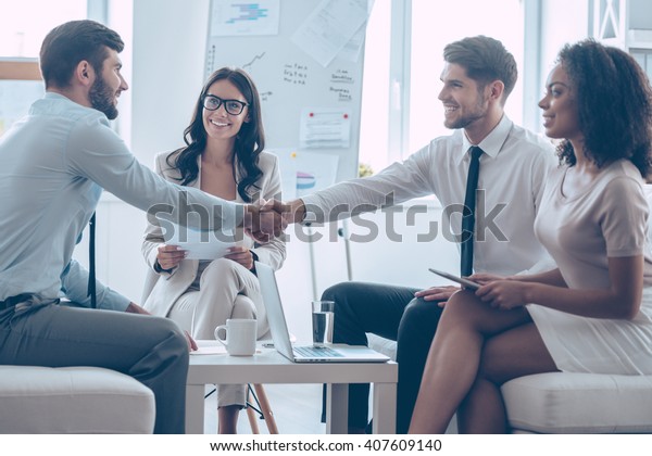 Welcome on\
board! Two handsome men shaking hands with smile while sitting on\
the couch at office with their\
coworkers