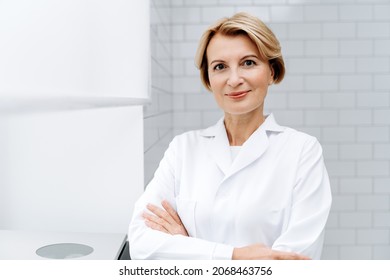 Welcome to our beauty salon. Portrait of mature cosmetologist in white lab coat looking at camera with smile while posing arms crossed at her clinic 