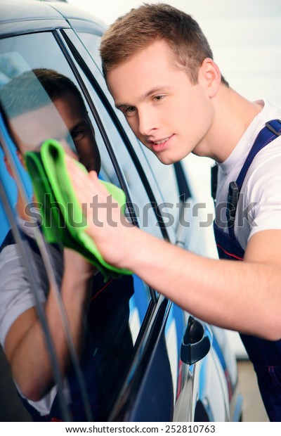 Welcome to our car\
service station. Closeup image of a handsome car mechanic wiping\
the car windows with tinting foil and smiling at camera in\
specialized service\
station