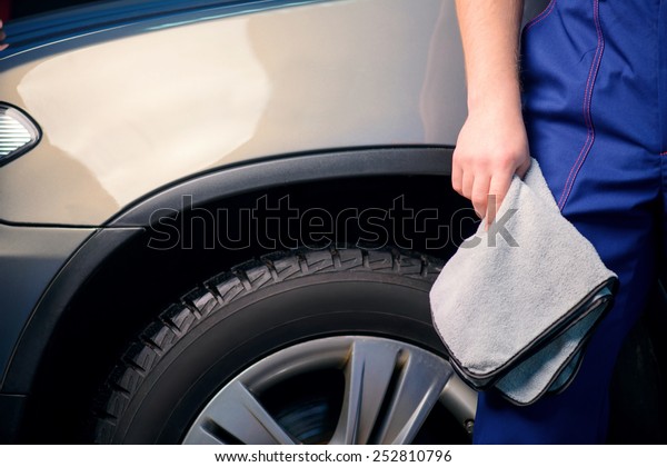 Welcome to our car service station. Closeup image\
of a handsome car mechanic in a uniform posing with a polishing\
wiper while standing against luxury suv in an authorized service\
station