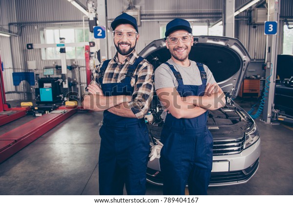 Welcome to our car service. Two experts  with
arms crossed, smiling at camera, in special safety outfit and
glasses, headwear, standing over background of entrance of
automobile in work
station