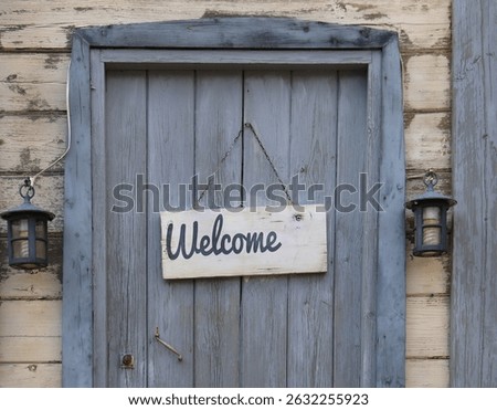 A welcome plank hanging in front of a rustic, wooden, closed door.