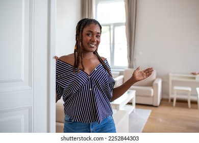 Welcome. Portrait of cheerful African woman inviting visitor to enter his home, happy young woman standing in doorway of modern apartment showing living room with hand