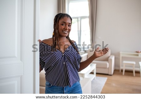 Welcome. Portrait of cheerful African woman inviting visitor to enter his home, happy young woman standing in doorway of modern apartment showing living room with hand