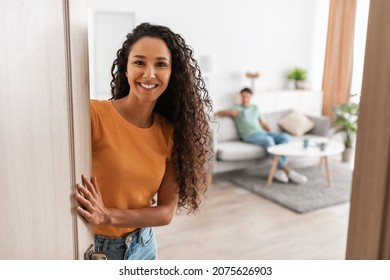 Welcome. Portrait of cheerful lady inviting guests to enter home, happy young lady standing in doorway of modern flat, holding door looking out, man sitting on couch in blurred background