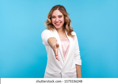 Welcome! Portrait of friendly hospitable cheerful woman with wavy hair in white jacket giving hand to handshake, hostess greeting guests, looking sociable positive. indoor studio shot, blue background - Shutterstock ID 1575536248