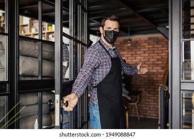 Welcome To The Restaurant, We Are Open. A Male Waiter In A Plaid Shirt And With A Black Apron Wears A Protective Face Mask And Greets Guests At The Entrance To The Facility And Opens The Door For Them