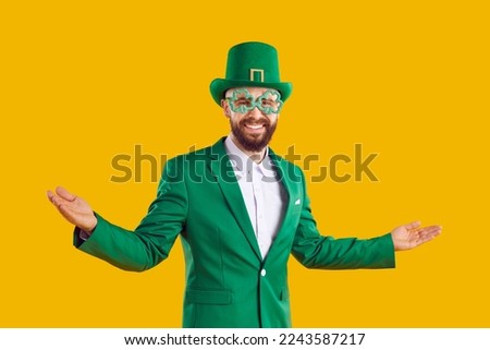Welcome to St Patrick's Day Party. Happy positive joyful cheerful bearded man wearing green suit, leprechaun hat and funny clover glasses smiling and spreading his hands on yellow studio background