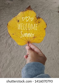 Welcome Summer Text On An Autumn Leaf In Yellow Color. A Young Woman Hand Holds The Leaf, White Sands Background. Human Body Part And Nature Photo Concept.