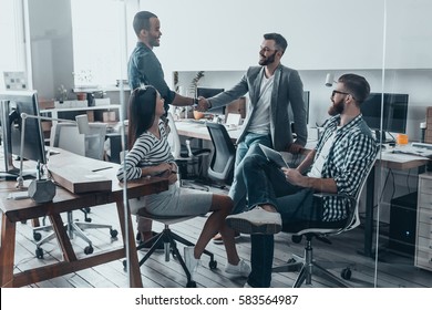 Welcome to team! Handsome young men in casual wear shaking hands and smiling while their colleagues looking at them and smiling - Powered by Shutterstock