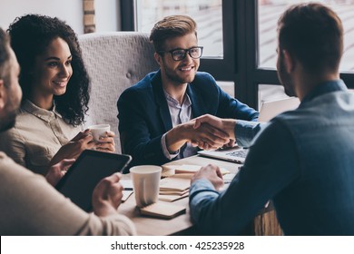 Welcome to team! Two men shaking hands and looking at each other with smile while their coworkers sitting at the business meeting 