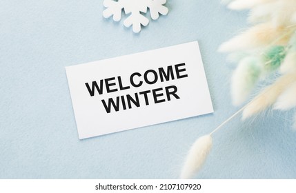 Welcome Winter Text On A Card On A Blue Background, Next To The Pathy Branches Of The Dried Flower And A Wooden Snowflake