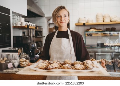 Welcoming female baker holding freshly baked almond croissants in  background of bakery. Young smiling seller woman in cafe or coffee shop. Bistro owner indoors. Sustainable local small business. - Powered by Shutterstock