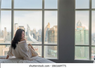 Welcoming a new day. Asian woman sitting on bed, entering a day happy and relaxed after good night sleep and city scenery in the window. Work-life balance city living quality concept. Copy space