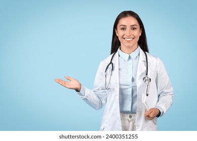 Welcoming young female doctor with a stethoscope makes an open hand gesture, as if introducing health services or explaining concepts, blue background - Powered by Shutterstock