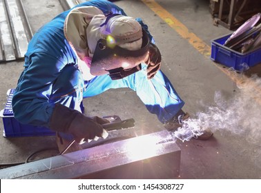 Welder is assembling the workpiece by process shielded metal arc welding (SMAW). Welder in blue uniform, safety shoes, leather gloves, welding mask. He is sitting and welding.