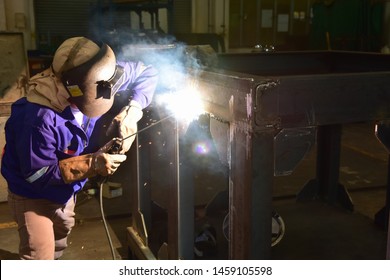 Welder is assembling the workpiece by process shielded metal arc welding (SMAW). Welder in blue uniform, safety shoes, leather gloves, welding mask. He is sitting and welding.