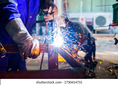 Welder is assembling the workpiece by process shielded metal arc welding (SMAW). Welder in blue uniform, safety shoes, leather gloves, welding mask. He is sitting and welding.