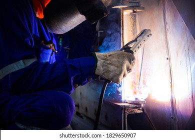 Welder is assembling the workpiece by process shielded metal arc welding (SMAW). Welder in blue uniform, safety shoes, leather gloves, welding mask. He is sitting and welding.