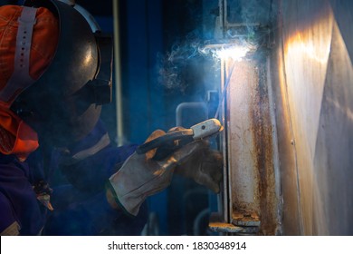 Welder is assembling the workpiece by process shielded metal arc welding (SMAW). Welder in blue uniform, safety shoes, leather gloves, welding mask. He is sitting and welding.