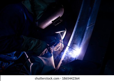 Welder is assembling the workpiece by process shielded metal arc welding (SMAW). Welder in blue uniform, safety shoes, leather gloves, welding mask. He working in the darkness.