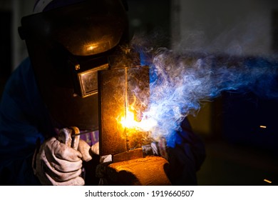 Welder is assembling the workpiece by process shielded metal arc welding (SMAW). Welder in blue uniform, safety shoes, leather gloves, welding mask. He is sitting and welding.