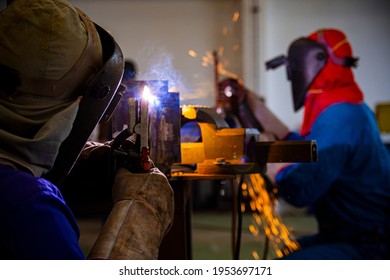 Welder is assembling the workpiece by process shielded metal arc welding (SMAW). Welder in blue uniform, safety shoes, leather gloves, welding mask. He is sitting and welding.