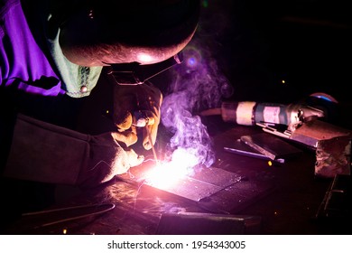 Welder is assembling the workpiece by process shielded metal arc welding (SMAW). Welder in blue uniform, safety shoes, leather gloves, welding mask. He is sitting and welding.