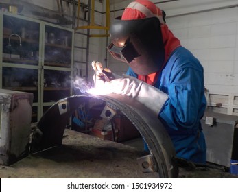 Welder is assembling the workpiece in workshop by process shielded metal arc welding (SMAW). Welder in blue uniform, safety shoes, leather gloves, welding mask. 